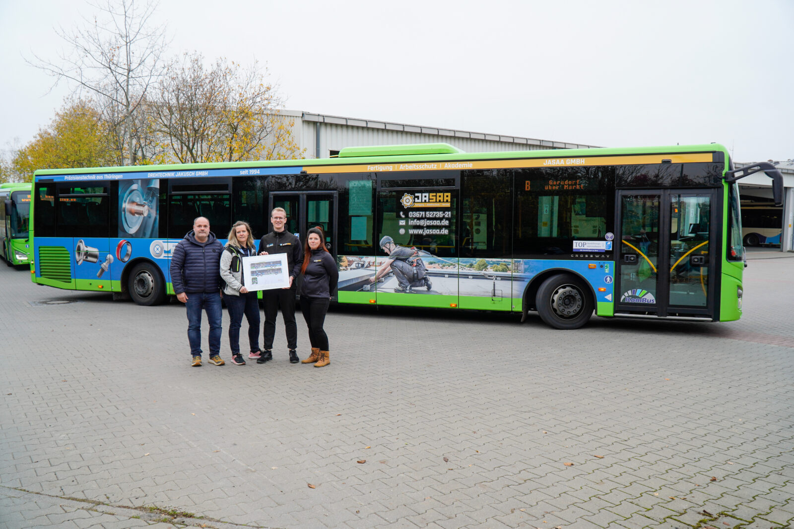 vier Personen stehen vor einem grün-blauen Bus mit einem Bild in der Hand 