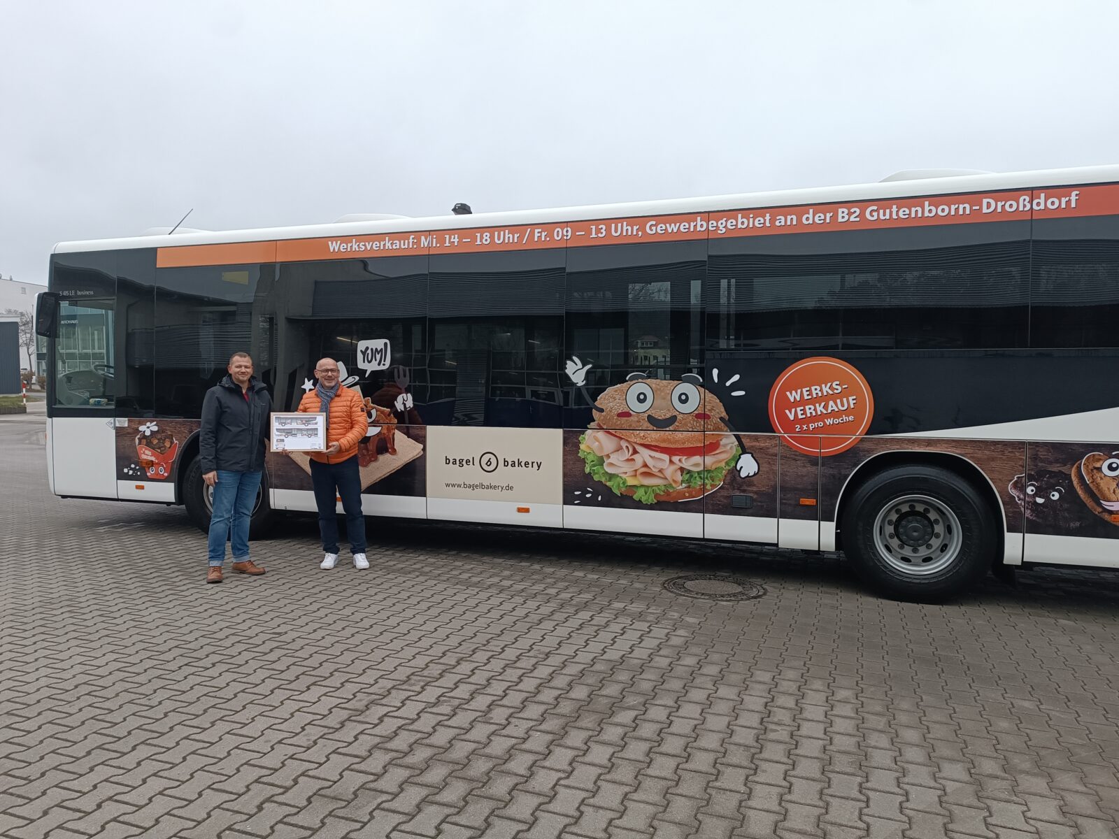 Zwei Männer stehen vor einem Bus auf dem ein Bagel zu sehen ist